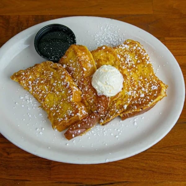 Golden French toast with powdered sugar, a scoop of whipped cream, and a side of syrup.