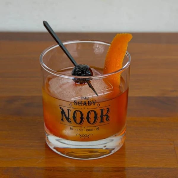 An Old Fashioned cocktail in a branded glass with an orange peel and cherry garnish.
