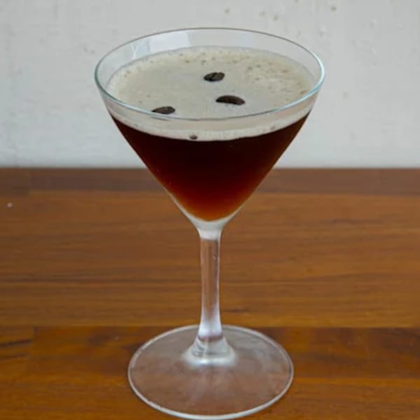 Espresso martini in a stemmed glass, topped with foam and three coffee beans on a wooden table.