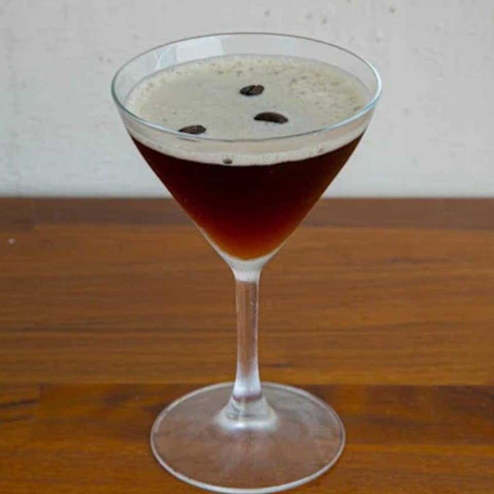 Espresso martini in a stemmed glass, topped with foam and three coffee beans on a wooden table.