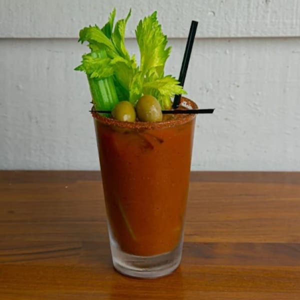 A Bloody Mary cocktail with celery, olives, and a black straw on a wooden table.